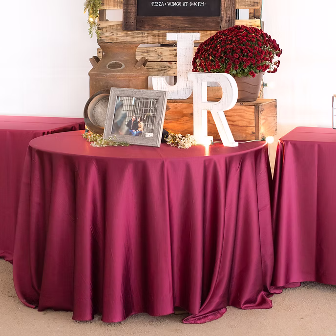 A round table with a burgundy tablecloth holds a framed photo, decorative flowers, large white wooden letters J and R, and rustic wooden boxes with a red potted plant, with candles and a sign in the background.