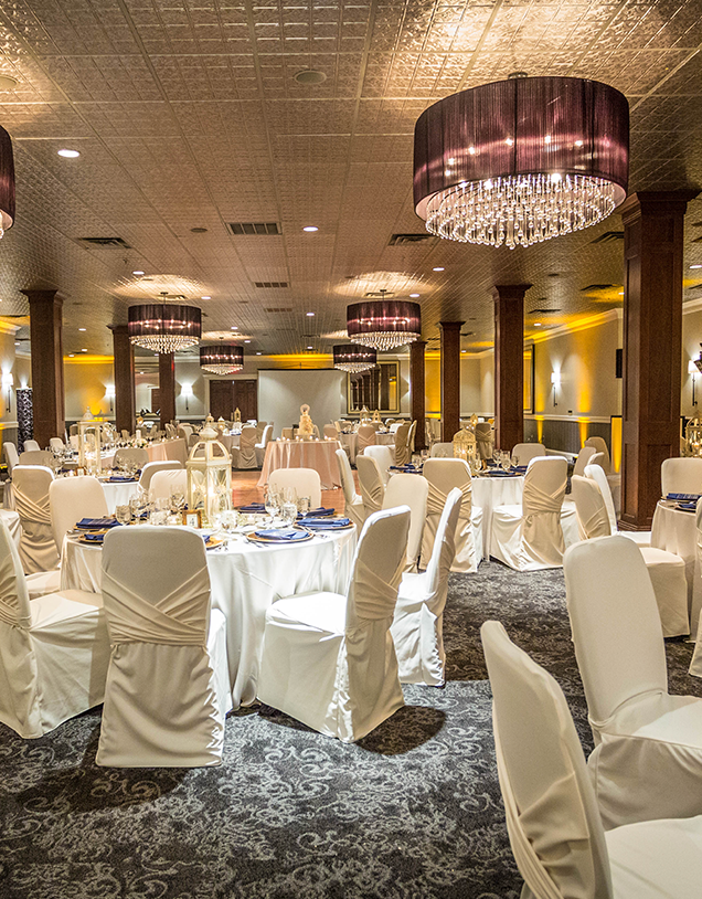 Elegant banquet hall with round tables covered in white cloths and chairs draped in white covers, set for an event. Large chandeliers hang from the ceiling, and warm lighting creates a festive atmosphere.