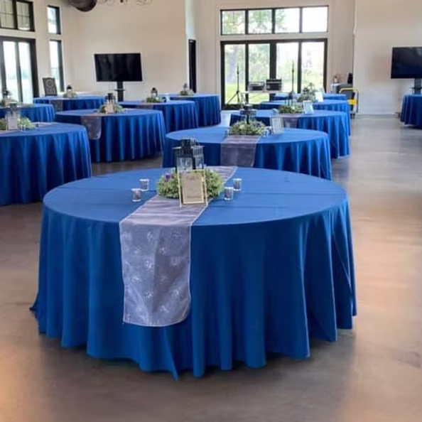 A spacious event hall with round tables covered in blue cloths and decorated with silver runners, centerpieces, and small candles. Large windows and TVs are visible in the background.