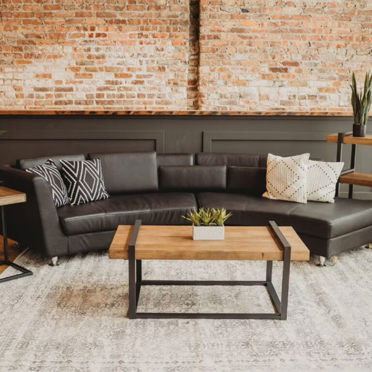 A modern living room with a black leather sectional sofa, patterned throw pillows, a wooden coffee table with a metal frame, a potted plant, and an exposed brick wall with black paneling behind it.
