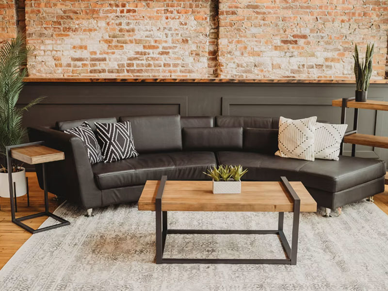 A modern living room with a black sectional sofa, patterned throw pillows, a wooden coffee table with black metal legs, a tall plant, and exposed brick walls. A light rug covers the wooden floor.