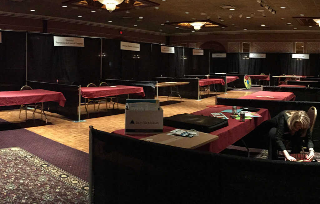 A large, dimly lit room with rows of empty tables covered in red tablecloths, informational signs above each station, and a person sitting alone at one table preparing materials.