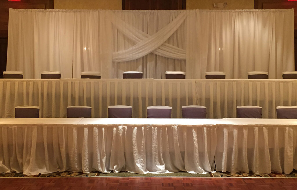A banquet hall setup with two long rows of tables draped in white cloths and chairs covered in white with purple sashes, in front of a white curtain backdrop with sheer fabric draped in an X shape.