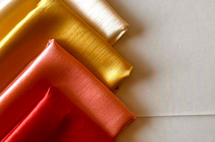 Four folded pieces of shiny fabric in cream, gold, orange, and red are arranged diagonally on a light, neutral background. The fabrics have a smooth, silky texture and reflective sheen.