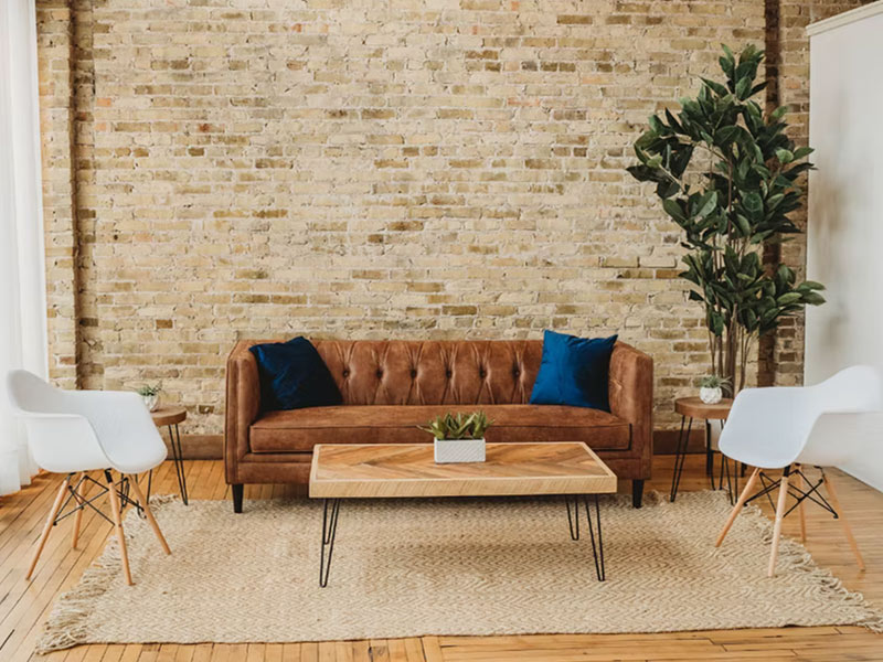 A modern living room with a brown leather sofa, two blue pillows, white accent chairs, a wooden coffee table, a green potted plant, and a beige rug on a wood floor against a brick wall.