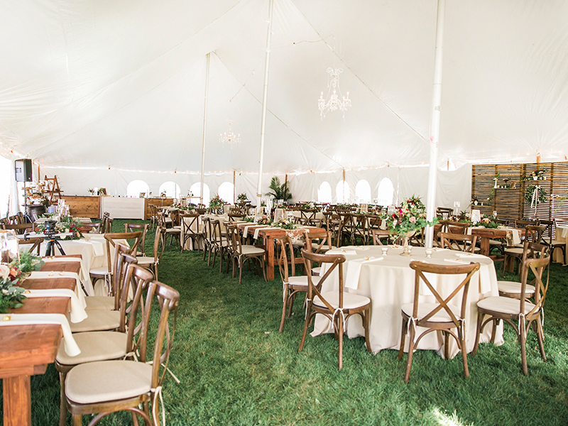 Elegant wedding reception setup under a large white tent with round tables, wooden chairs, flower centerpieces, chandeliers, and green grass flooring. Natural light fills the spacious, inviting atmosphere.