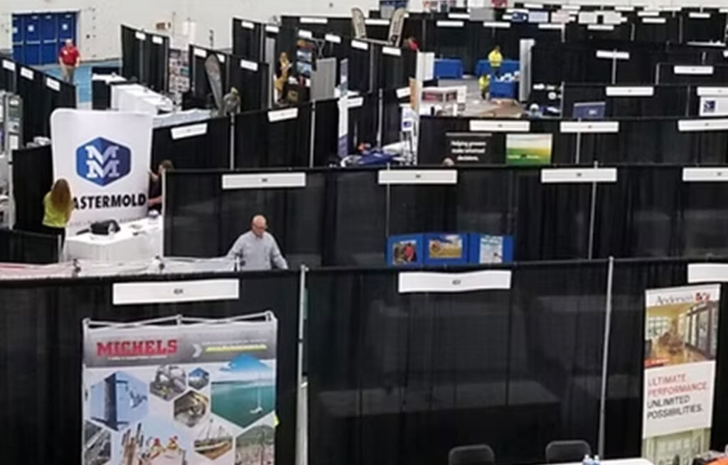 A trade show or convention floor with multiple black booths, banners, and displays. Some booths have company logos and posters. A few people are walking and setting up in the aisles.