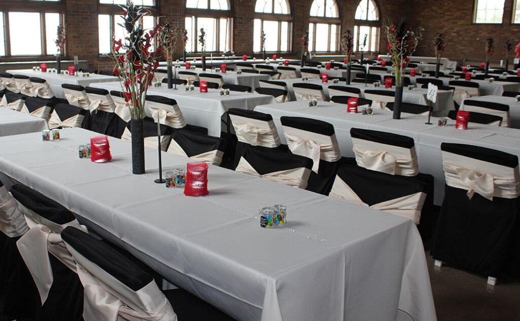 Rows of banquet tables covered with white tablecloths, black chair covers with white sashes, red and black centerpieces, and small glass decorations, set in a large room with arched windows and brick walls.