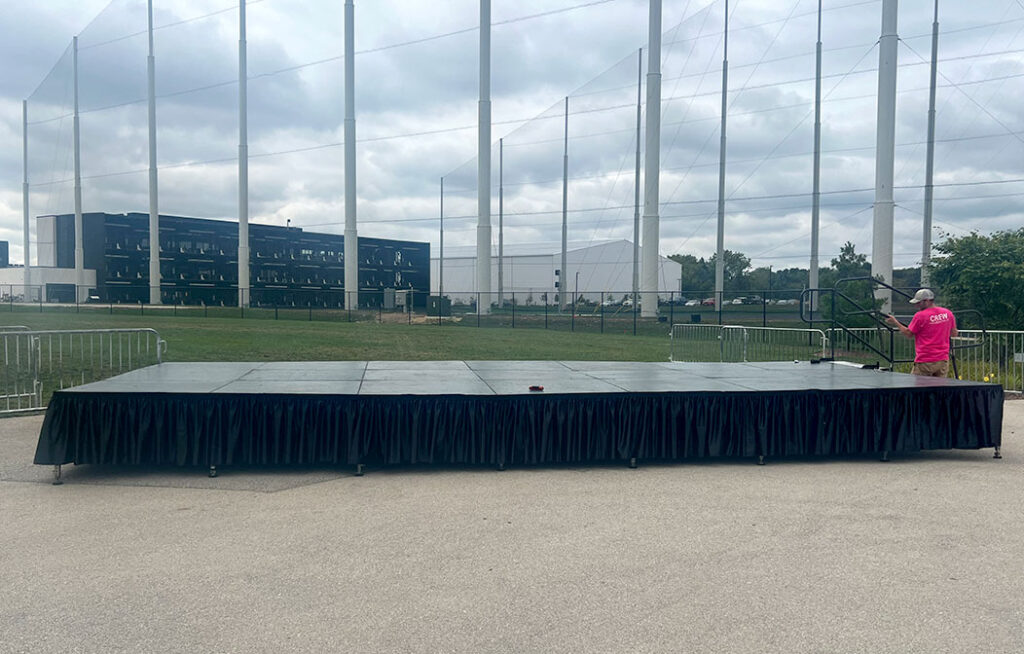 An empty outdoor stage with a black skirt, set up on pavement, with a person in a pink shirt standing to the right. Fenced green grass, netted poles, and a building are visible in the background under a cloudy sky.
