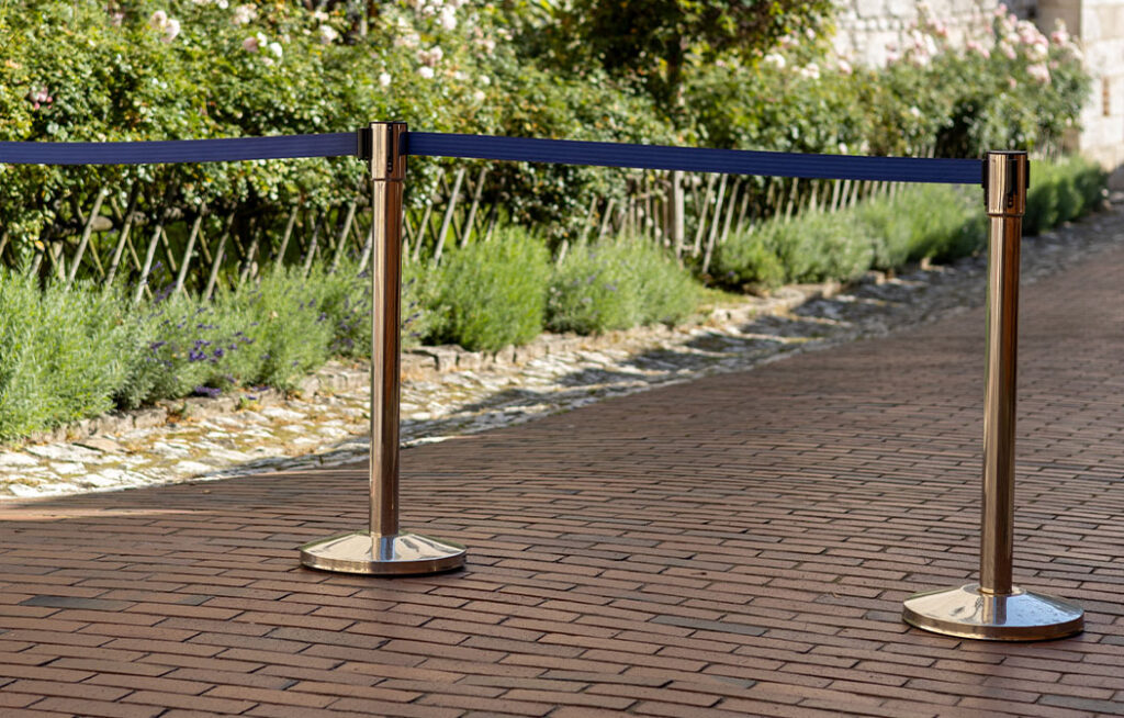 Two stanchions with a blue rope block off a section of a brick pathway next to a garden with green plants and flowers.