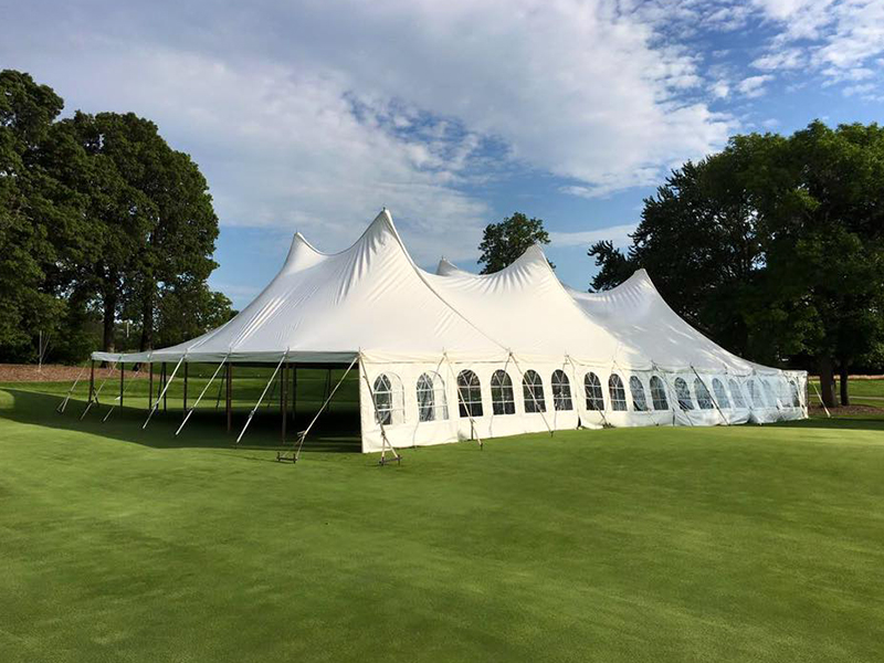 A large white event tent with arched windows is set up on green grass, surrounded by trees under a partly cloudy sky.