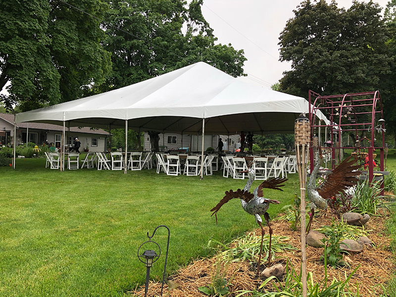 A large white event tent with tables and chairs set up underneath is on a green lawn, surrounded by trees and garden decorations, including metal crane sculptures and a red arbor.