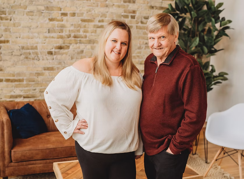 A woman in a white off-the-shoulder top stands next to an older man in a red sweater. They are smiling, indoors, with a couch, plant, and brick wall in the background.