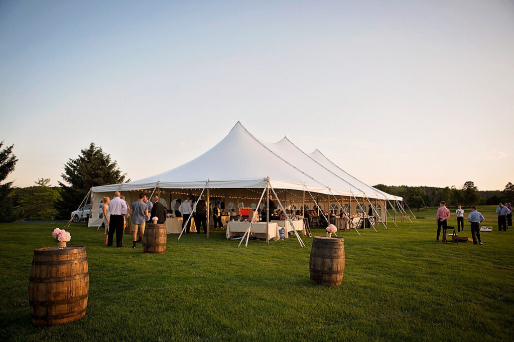 A large white event tent is set up on a grassy lawn with people gathered around outside. Wooden barrels and small tables are scattered on the grass, and the sky is clear at sunset.