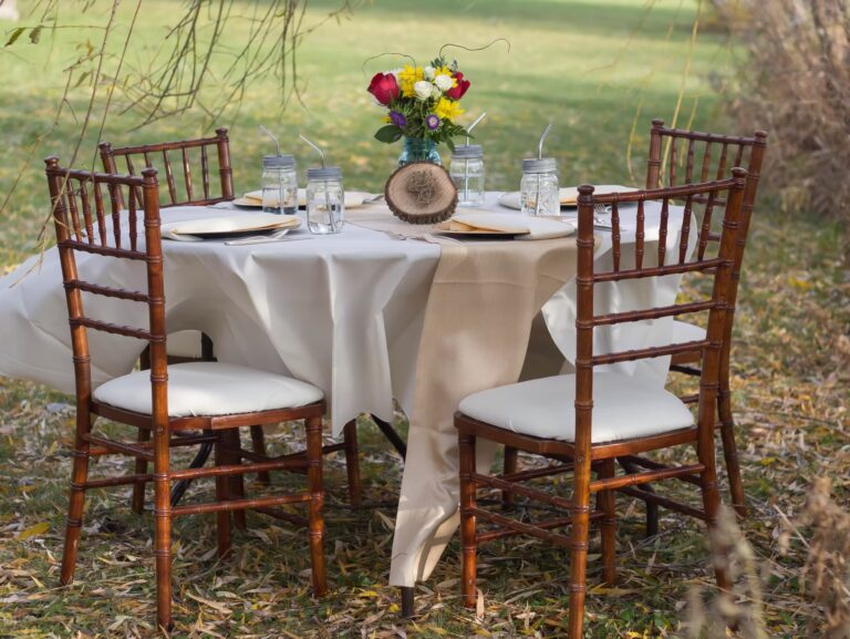 A round outdoor table set for four with wooden chairs, white tablecloth, beige runner, mason jar glasses with straws, plates, and a colorful flower centerpiece, surrounded by grass and autumn leaves.