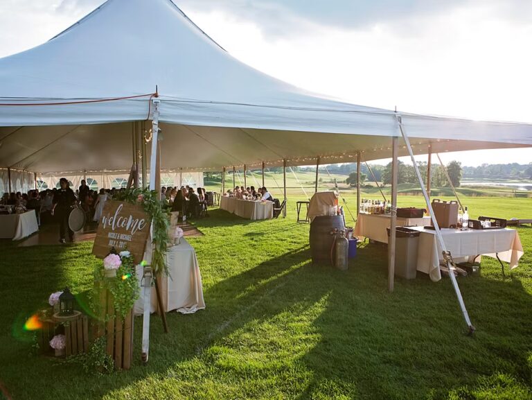 A large white tent set up on grass for an outdoor event, with guests seated at tables inside. A wooden welcome sign stands at the entrance, and a beverage station is set up outside. Sunlight shines on the green lawn.