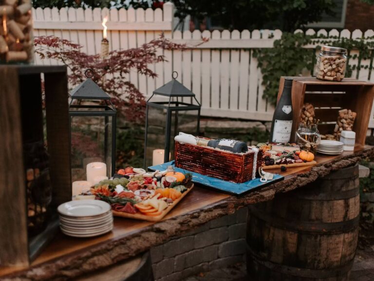 A rustic outdoor charcuterie setup on a wooden plank atop barrels features assorted meats, cheeses, fruits, stacked plates, candles, wine, glass jars filled with corks, and lanterns, against a white picket fence backdrop.