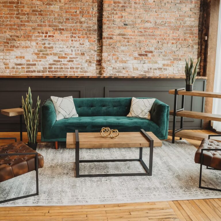 A stylish living room with a green velvet sofa, two geometric pillows, wooden coffee table, brown tufted ottomans, metal shelves, potted plants, and an exposed brick wall background.