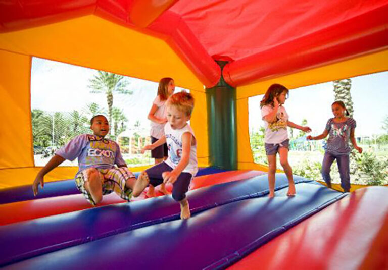 Four children play and jump together inside a colorful inflatable bounce house, with sunlight and palm trees visible through the window openings in the background.