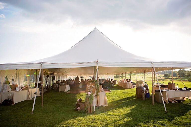 A large white event tent set up outdoors on green grass, filled with people seated at tables, with sunlight streaming in and decorations around the entrance.