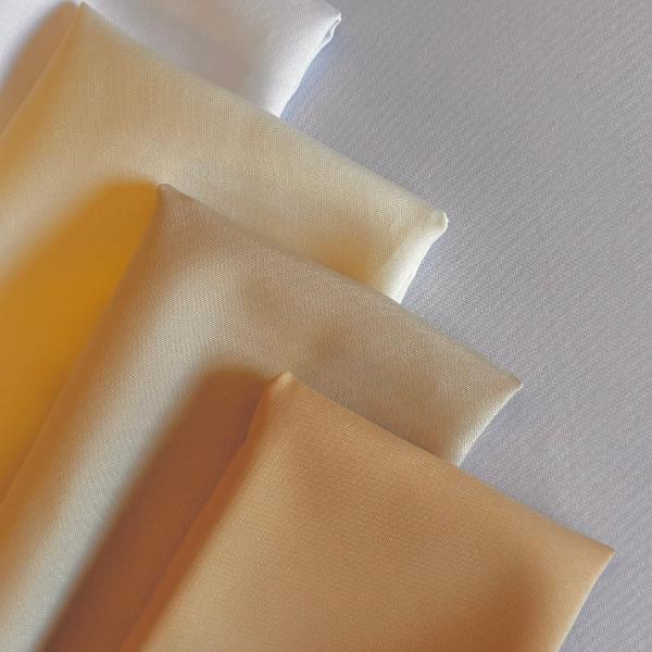 Four folded fabric pieces in soft, neutral colors—white, ivory, beige, and light tan—are arranged in parallel on a textured white background, showing their smooth, matte surfaces and subtle color differences.