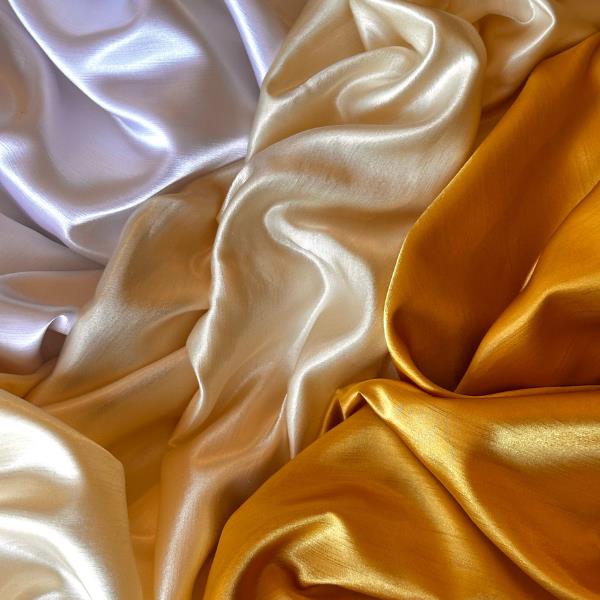 Shiny, smooth fabric in three colors—white, cream, and golden yellow—draped in soft, flowing folds, creating a luxurious and elegant texture.