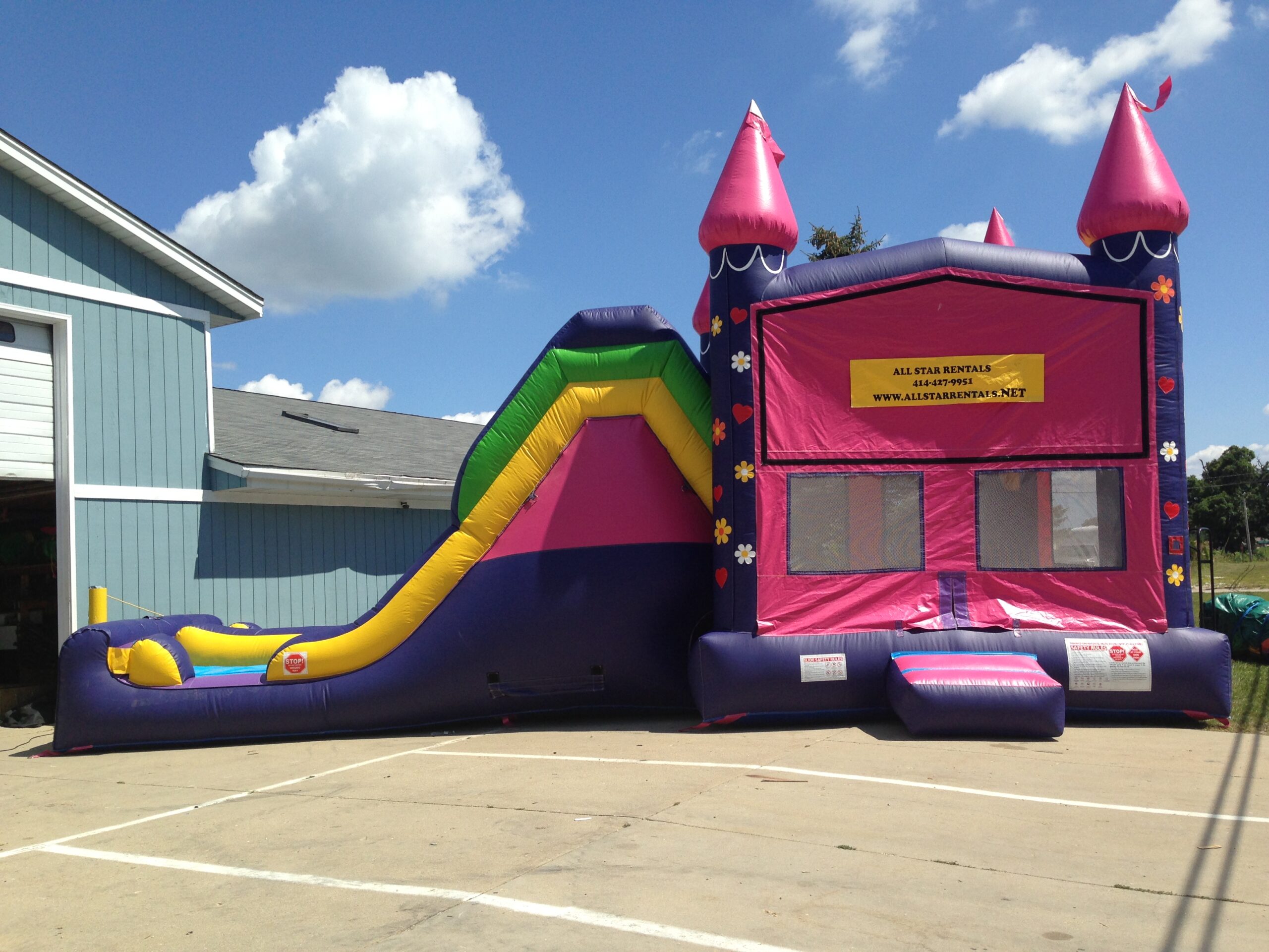 A pink and purple inflatable bounce house with castle towers is connected to a slide. The setup is outdoors on a sunny day, next to a light blue building. A yellow sign with rental information is on the bounce house.