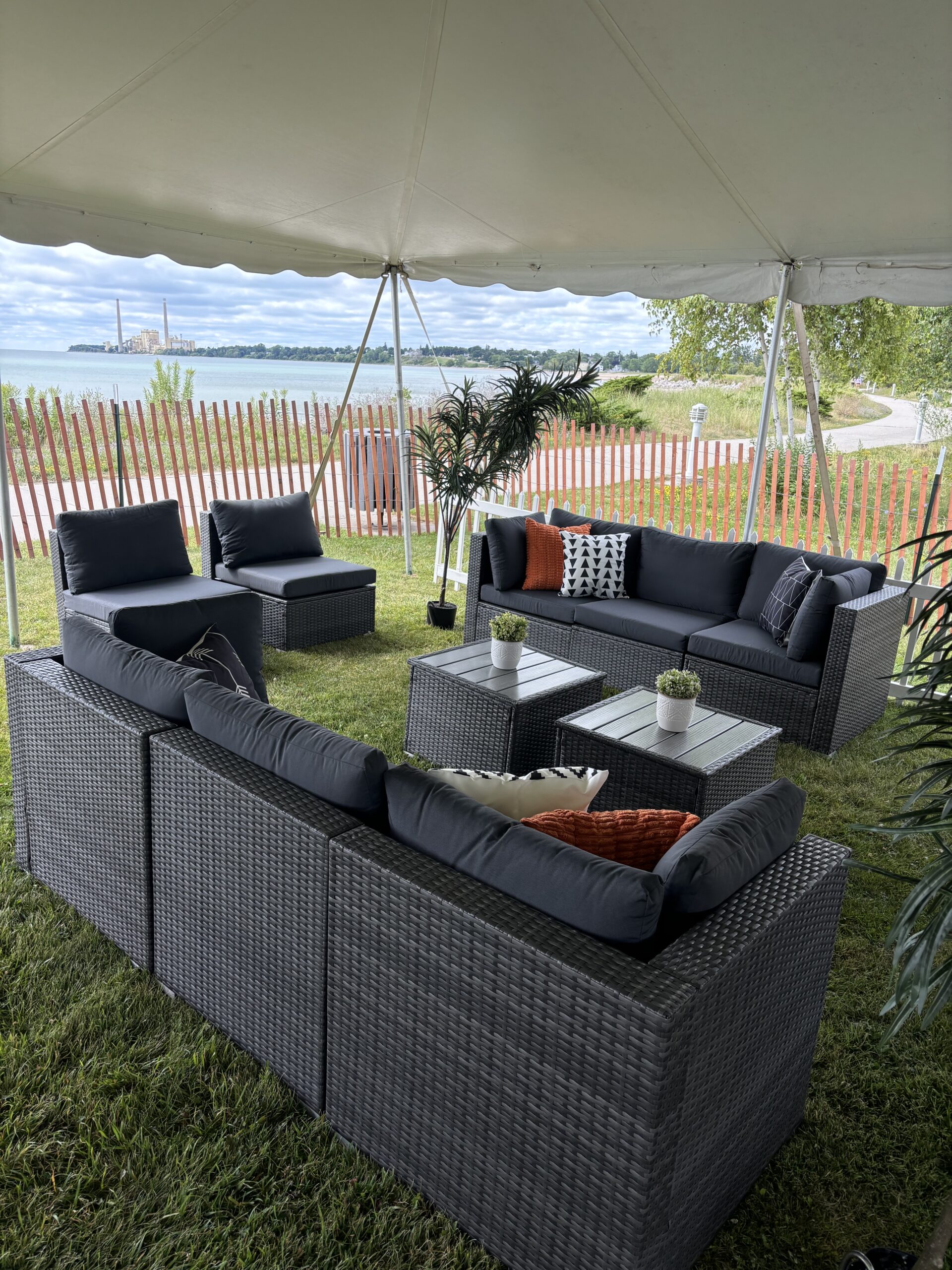Outdoor lounge area under a tent with gray wicker sofas, black cushions, and small coffee tables with potted plants. The setup overlooks water, a wooden fence, and a distant industrial landscape.