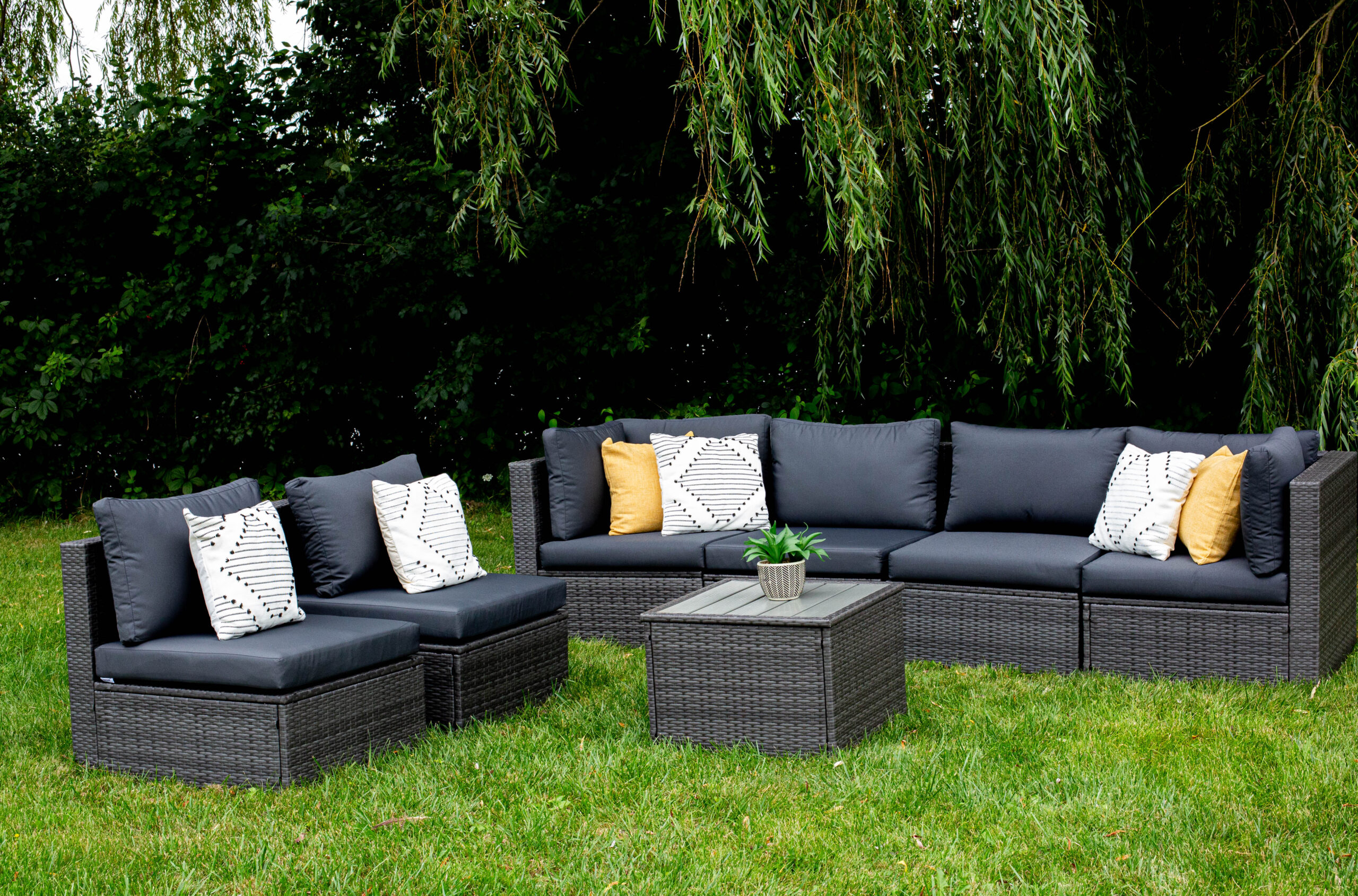 Outdoor patio set with dark wicker furniture and navy cushions, arranged in an L-shape on a lawn. Decorative pillows in white patterns and yellow add color, with a potted plant on the square coffee table. Trees provide shade in the background.