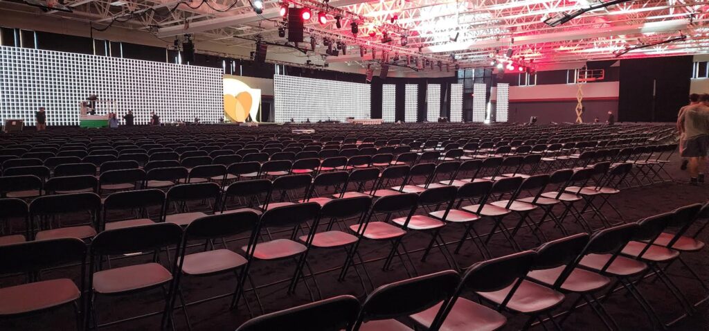 Rows of empty black chairs are arranged in a large indoor venue with a patterned backdrop, stage, bright overhead lights, and a few people preparing for an event.