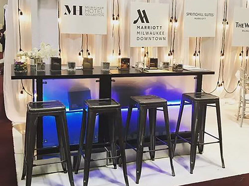 A modern bar-height table with four metal stools in front of a white curtain backdrop, featuring signs for different hotels and soft string lighting. The table is lit with blue accent lights underneath.