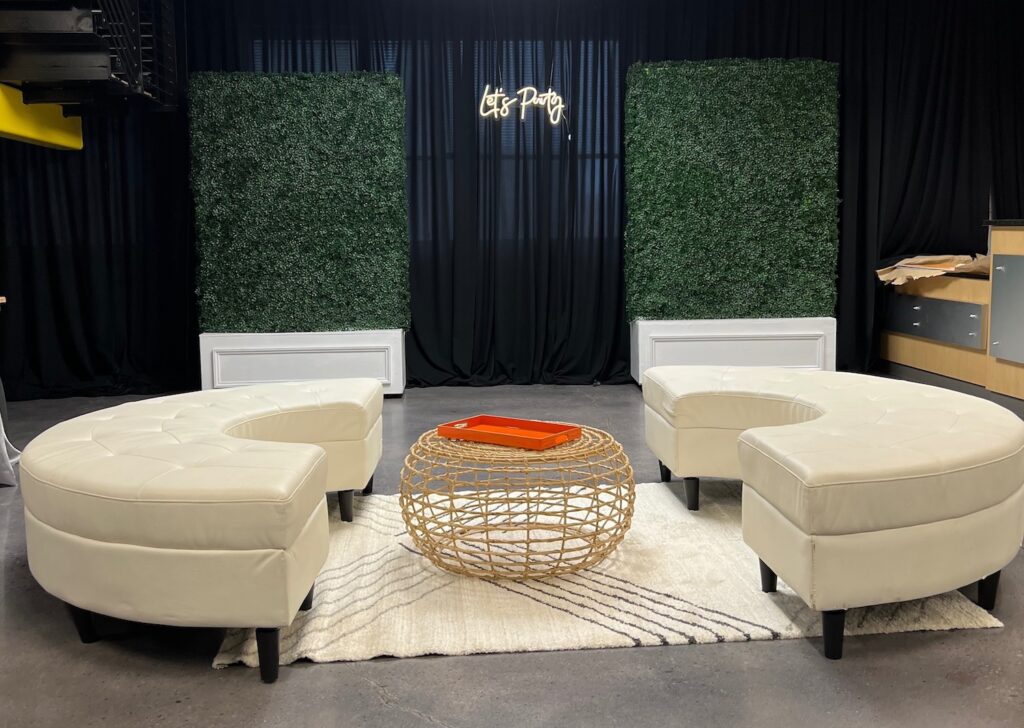 Modern lounge setup with two curved white leather sofas facing each other, a round woven coffee table with an orange tray, white rug, artificial hedge panels, and a Lets Party neon sign against black curtains.