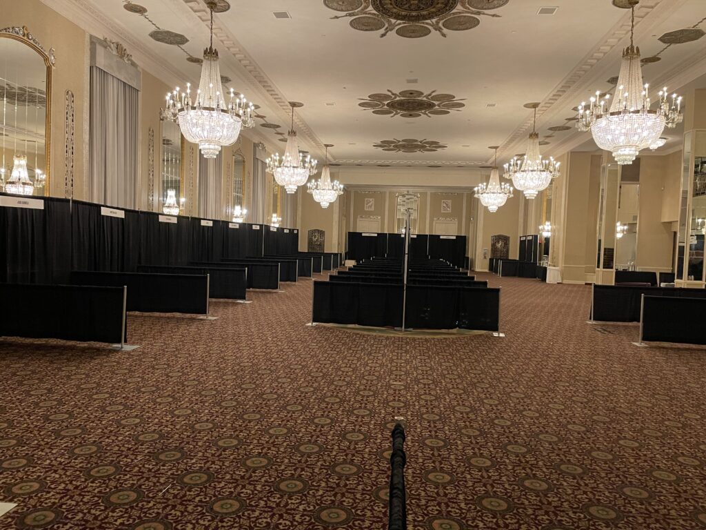 A large, elegant ballroom with ornate chandeliers, patterned carpet, high ceilings, and black curtains forming partitioned booths. The room is empty and well-lit, with mirrors and decorative details on the walls.