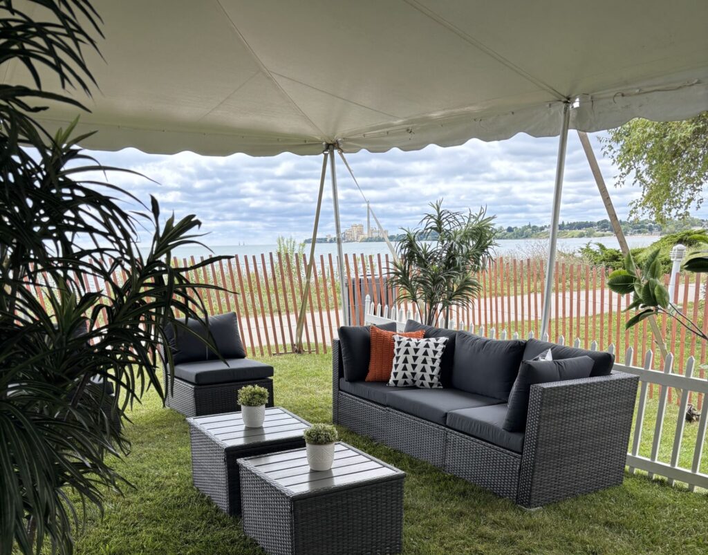 Outdoor seating area under a tent with wicker sofas and chairs, dark cushions, small tables, potted plants, and a grassy lawn. A white fence and scenic view with cloudy sky appear in the background.