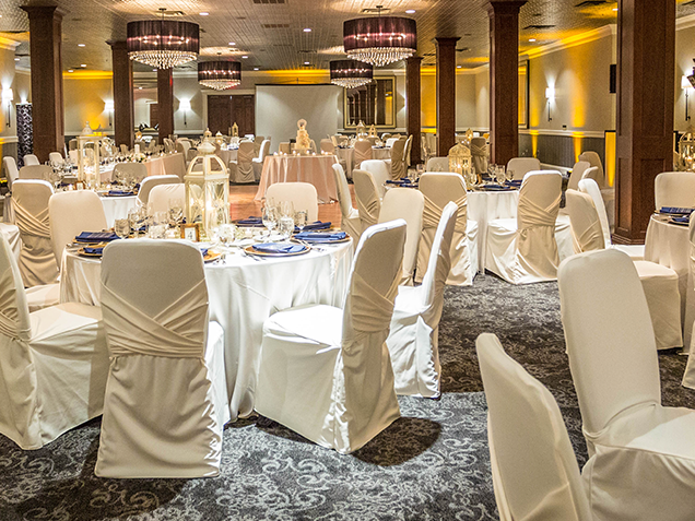 Elegant banquet hall with round tables covered in white cloths and chairs draped in white covers, set for an event. Large chandeliers hang from the ceiling, and warm lighting creates a festive atmosphere.