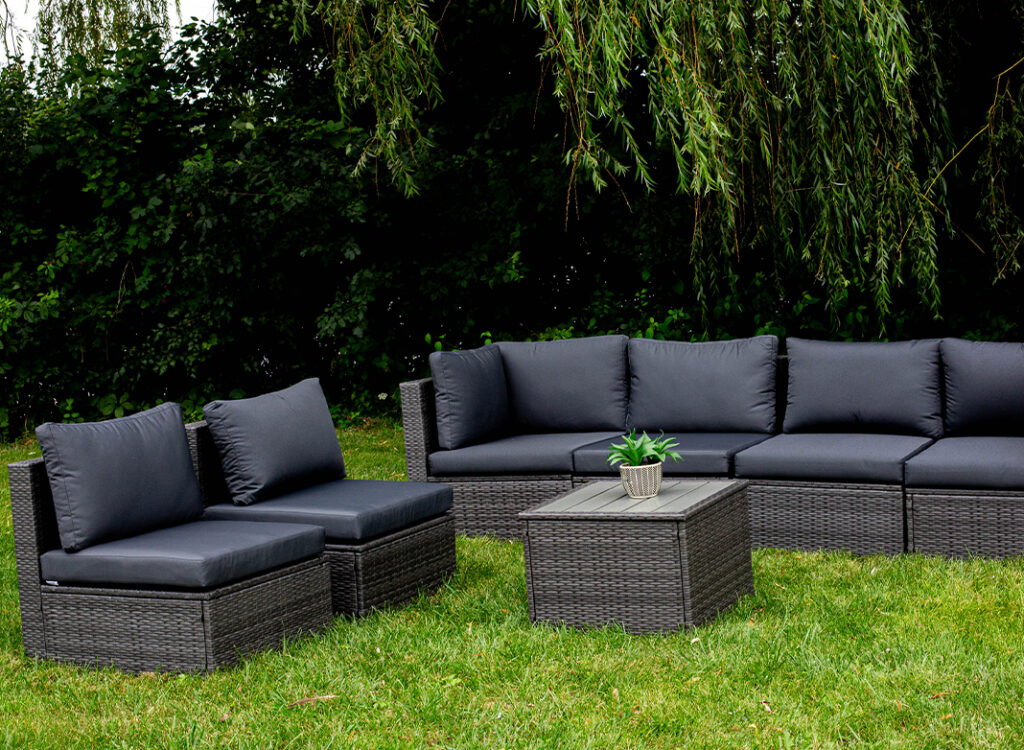 Outdoor wicker patio furniture set with black cushions, including a sectional sofa, two chairs, and a square coffee table topped with a small potted plant, placed on green grass with trees in the background.