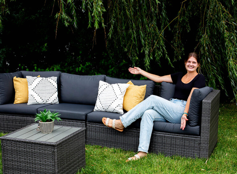 A woman sits smiling on a gray outdoor sectional sofa with patterned and yellow pillows, gesturing towards the seating. The sofa is on grass beside a small table with a potted plant, under leafy trees.