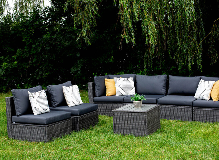 Outdoor patio set with dark rattan furniture, featuring gray cushions, patterned and yellow throw pillows, and a square coffee table with a small potted plant on top, set on green grass under hanging tree branches.