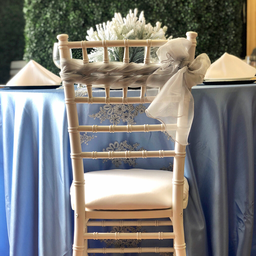 Beyond folding chairs: Elevate your corporate event with stylish furniture like a white wooden chair adorned with a sheer gray ribbon, set before a round table dressed in light blue and accented by elegant napkins and floral decor.