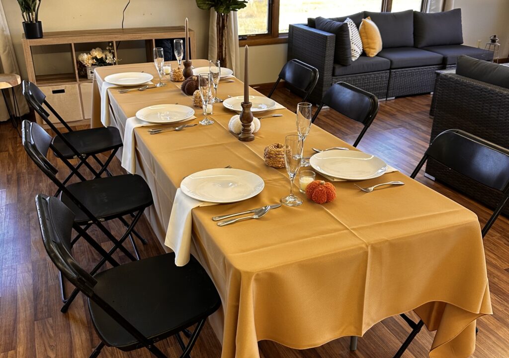 A dining table with a yellow tablecloth is set for six with white plates, silverware, and glasses. Small decorative pumpkins and vases are placed in the center. Rent tables, linens, and chairs for your holiday party to create this cozy look.