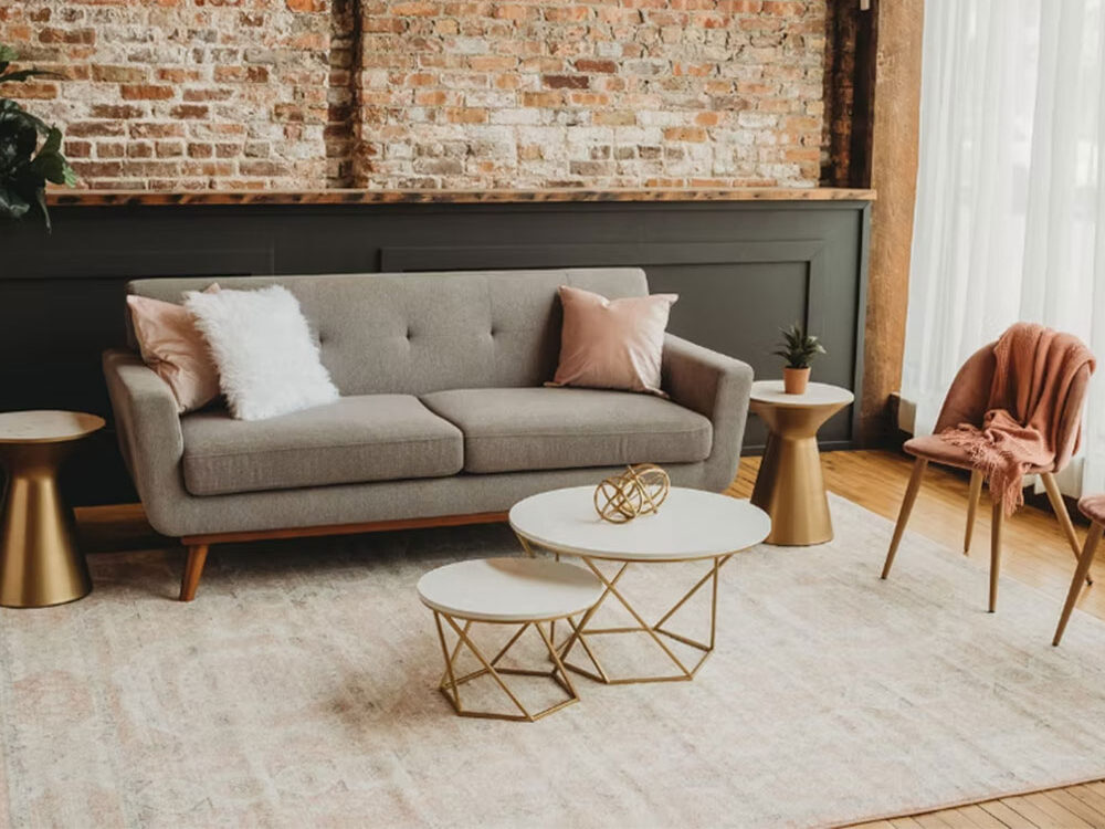 A modern living room with a grey sofa, pink and white pillows, a pink throw on a chair, gold side tables, geometric coffee tables, a plant, brick wall, and large sheer curtains letting in natural light.