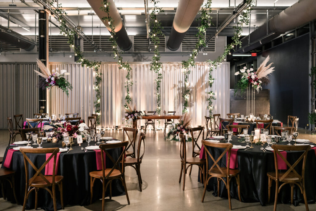 A modern, elegant event space set for a formal dinner with round tables draped in black tablecloths, wooden chairs, floral centerpieces, hanging greenery, and soft lighting. A sweetheart table is featured at the back under an arch.