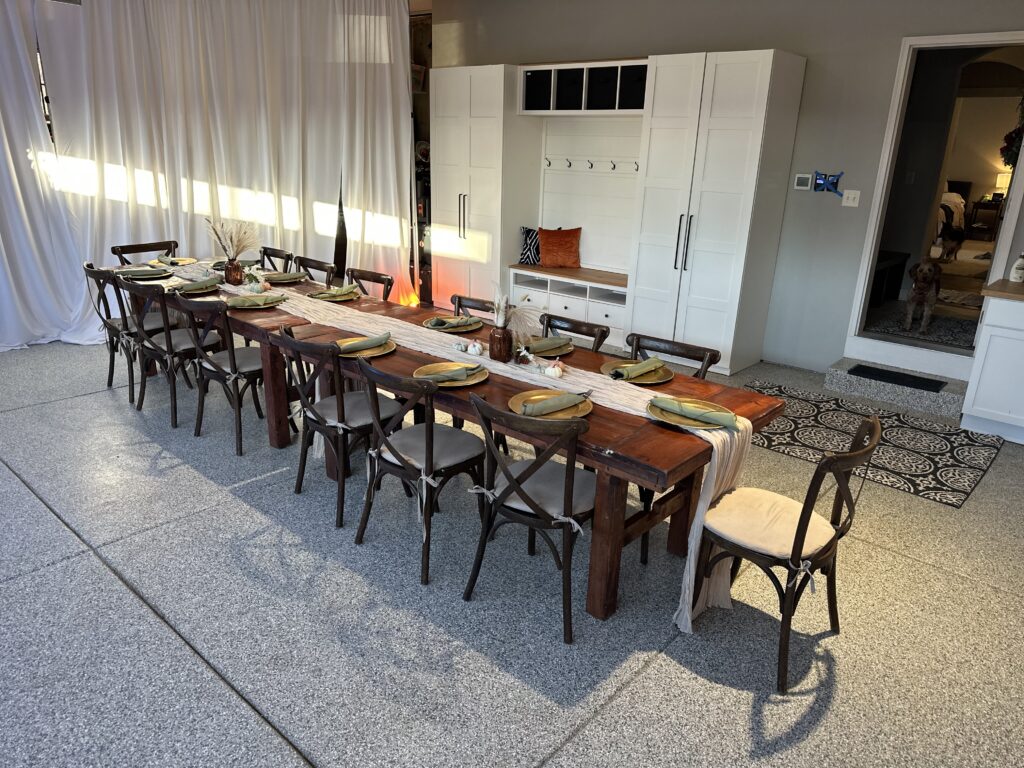 A long wooden dining table with twelve chairs is set for a meal, featuring gold plates and neutral runners—beyond folding chairs: elevate your corporate event with stylish furniture. A white cabinet stands against the wall, and a dog is near a doorway in the background.