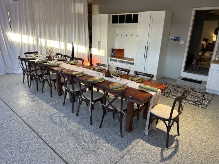 A long wooden dining table with twelve chairs is set for a meal, featuring gold plates and neutral runners—beyond folding chairs: elevate your corporate event with stylish furniture. A white cabinet stands against the wall, and a dog is near a doorway in the background.