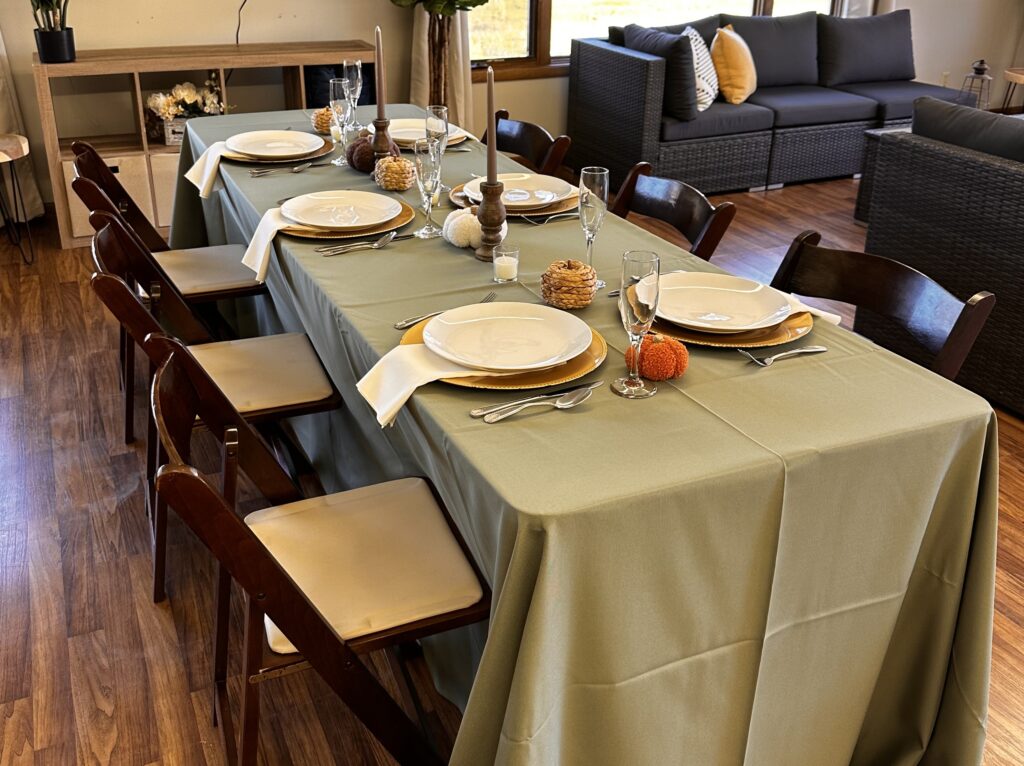 A dining table set for six with a light green tablecloth, white plates, silver cutlery, and ivory napkins—rent tables, linens, and chairs for your holiday party to recreate this cozy look with pumpkins and gourds as centerpieces.
