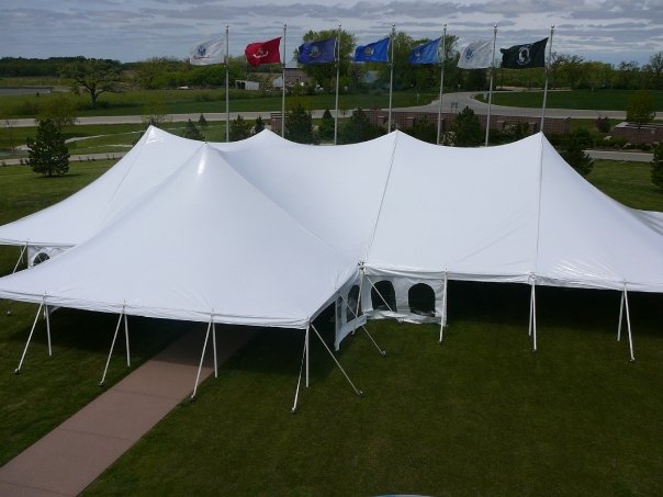 Large white event tents are set up on a green lawn with eight flags flying in the background, making this space perfect for outdoor gatherings and inspiring holiday party ideas. A paved walkway winds beside the tents with trees and a road in the distance.
