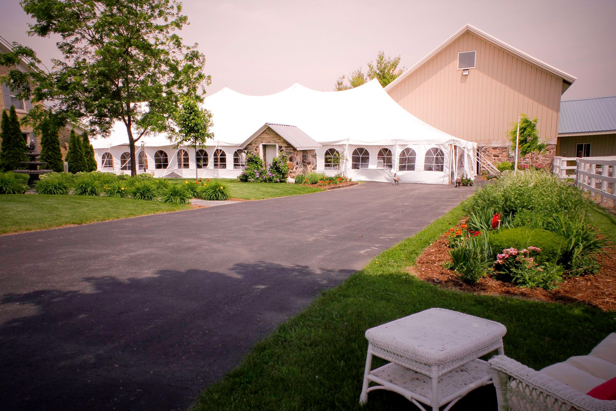 A large white event tent with arched windows is set up beside a beige barn, perfect for gathering holiday party ideas. The scene features a paved driveway, green lawn, flower beds, and white wicker patio furniture in the foreground.