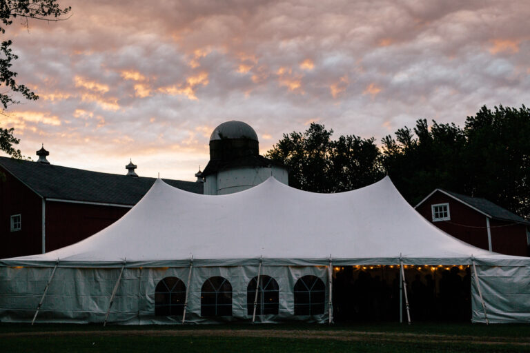 A large white event tent stands in front of a red barn and trees at sunset, string lights glowing inside. With dramatic clouds above, it’s an inspiring backdrop for holiday party ideas.