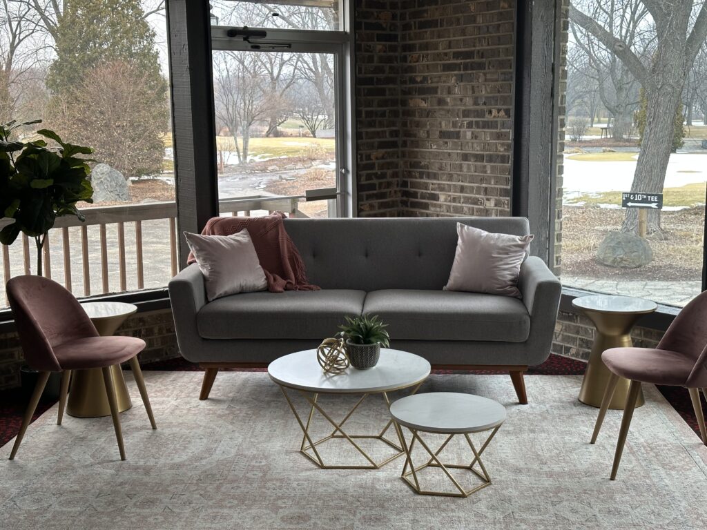 A modern living room with a gray sofa, pink pillows, and a throw blanket—perfect for holiday party ideas. Two pink chairs with gold bases, marble-top tables, a potted plant, and large windows offer a cozy view of the wintery scene outside.