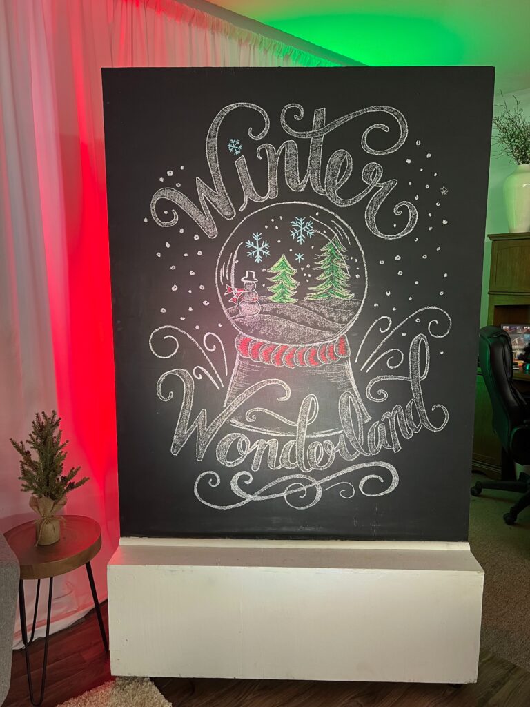 A black chalkboard displays Winter Wonderland in decorative letters with a snow globe drawing of trees and a snowman. The room, lit with red and green lights, inspires festive holiday party ideas and evokes a cheerful holiday atmosphere.